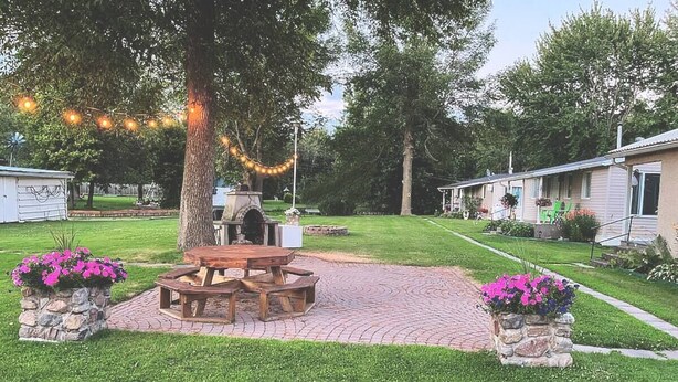 Un espace pique-nique avec une table et des bancs en bois, entouré de jardinières en pierre ornées de fleurs, sous un grand arbre illuminé de guirlandes lumineuses.