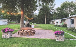 Un área de picnic con una mesa de madera y bancos, rodeada de macetas de piedra con flores, bajo un gran árbol con luces colgantes.