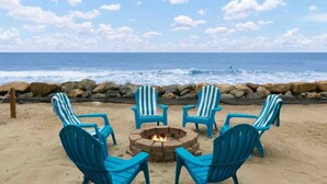 Une plage avec quatre chaises Adirondack bleues disposées autour d’un foyer en pierre, offrant une vue sur l’océan.