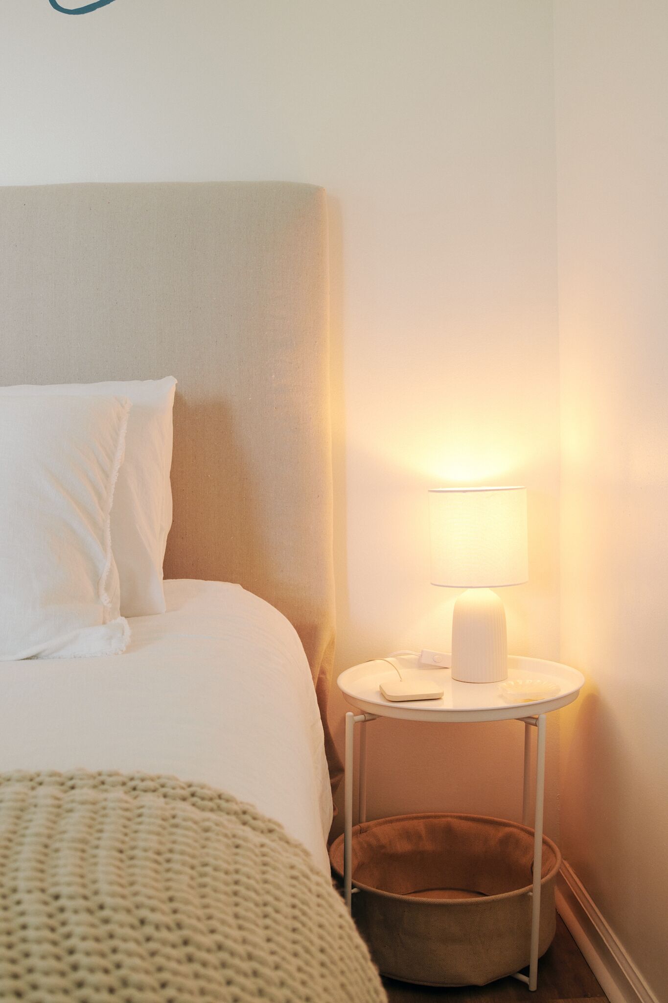 A neatly made bed with white linens, a bedside table with a lamp, and a small storage basket.