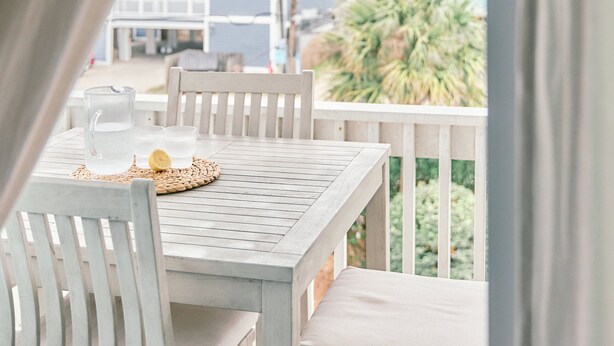 Une table avec une carafe, des verres et un citron, le tout posé sur une nappe blanche, sur un balcon doté de balustrades blanches et offrant une vue sur des immeubles et des palmiers.