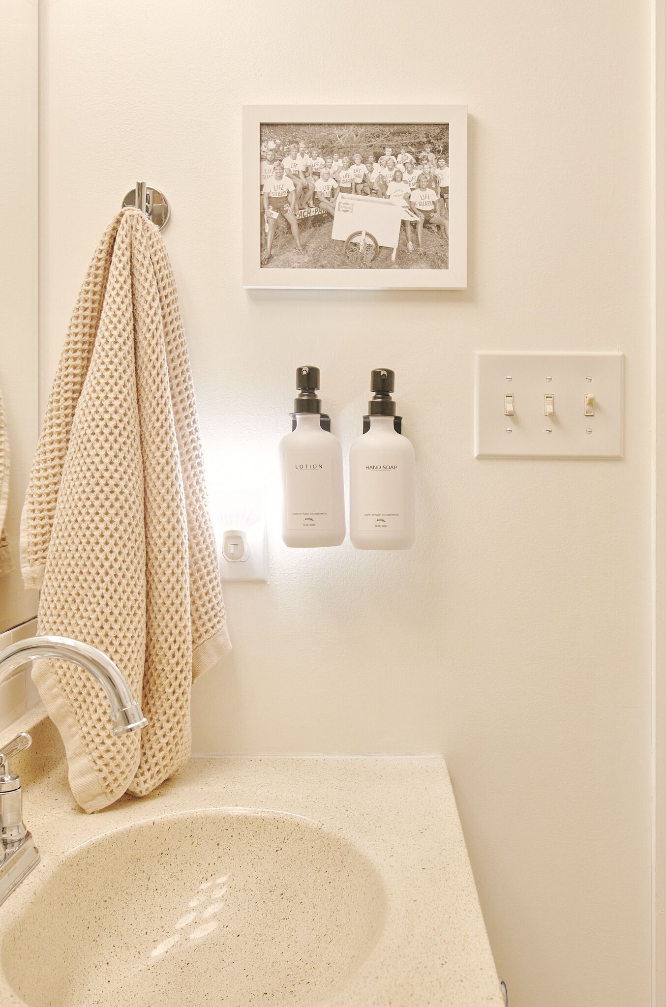 Un bagno con un lavandino color crema, un porta asciugamani con un asciugamano a pois, una cornice con una foto appesa al muro e due dispenser per sapone liquido.