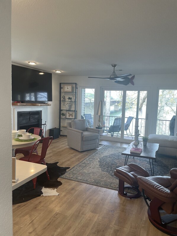 A spacious living room with a fireplace, a dining area with a table and chairs, a ceiling fan, and large windows offering a view of the outdoors.