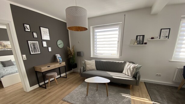 Ett modernt vardagsrum med en grå soffa, ett skrivbord i trä, ett vitt soffbord och ett stort fönster med persienner.