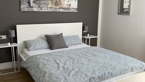 Ett modernt sovrum med en vit säng, grått och vitt mönstrat sänglinne, en grå kudde, ett litet sängbord med en lampa och en abstrakt tavla som hänger på väggen.