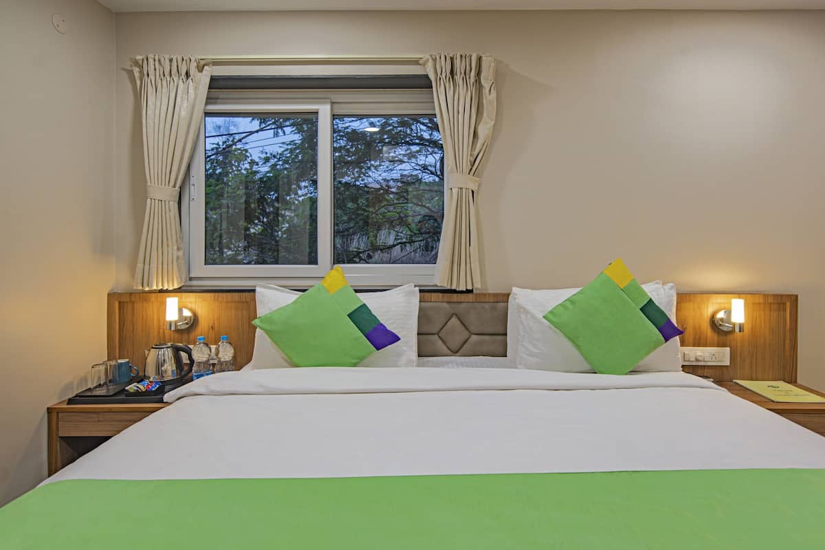 Quarto de hotel com cama, duas mesas de cabeceira, uma janela com cortinas e vista para árvores.