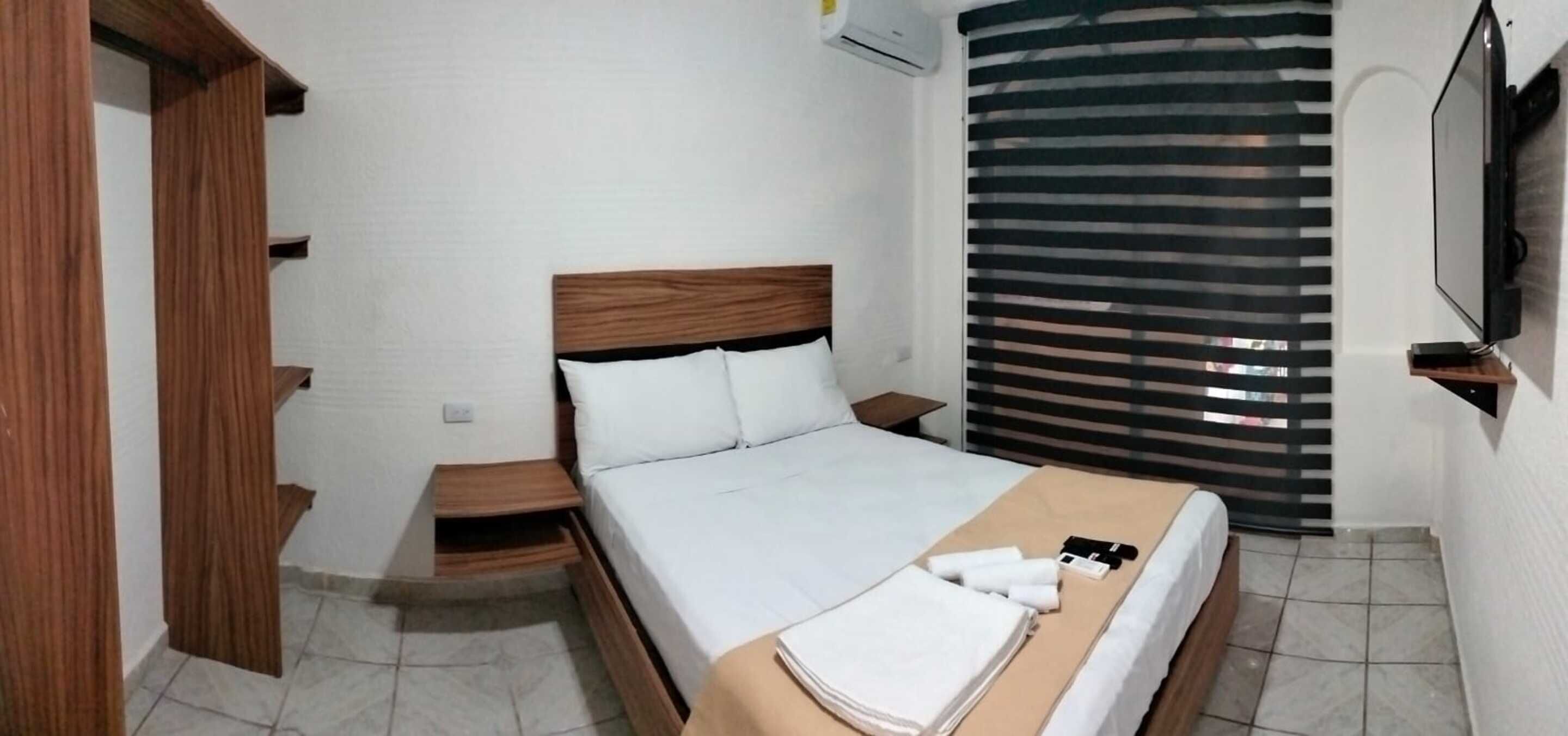 A hotel room with a bed, a nightstand, a wooden wardrobe, a TV, and a window with blinds.