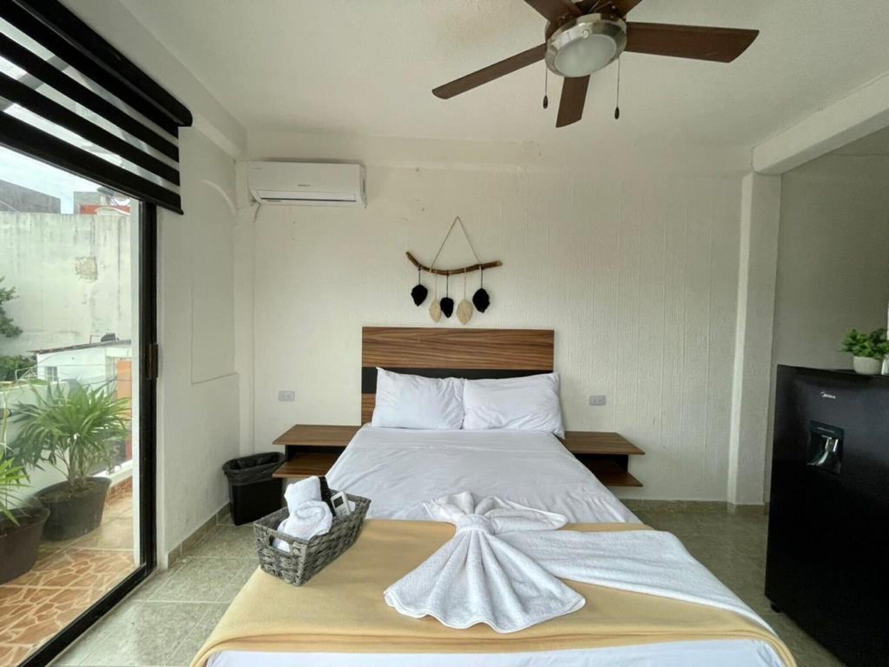 Hotelkamer met een bed, een plafondventilator, een wandairconditioner, een klein nachtkastje met een mand, en uitzicht op buiten.