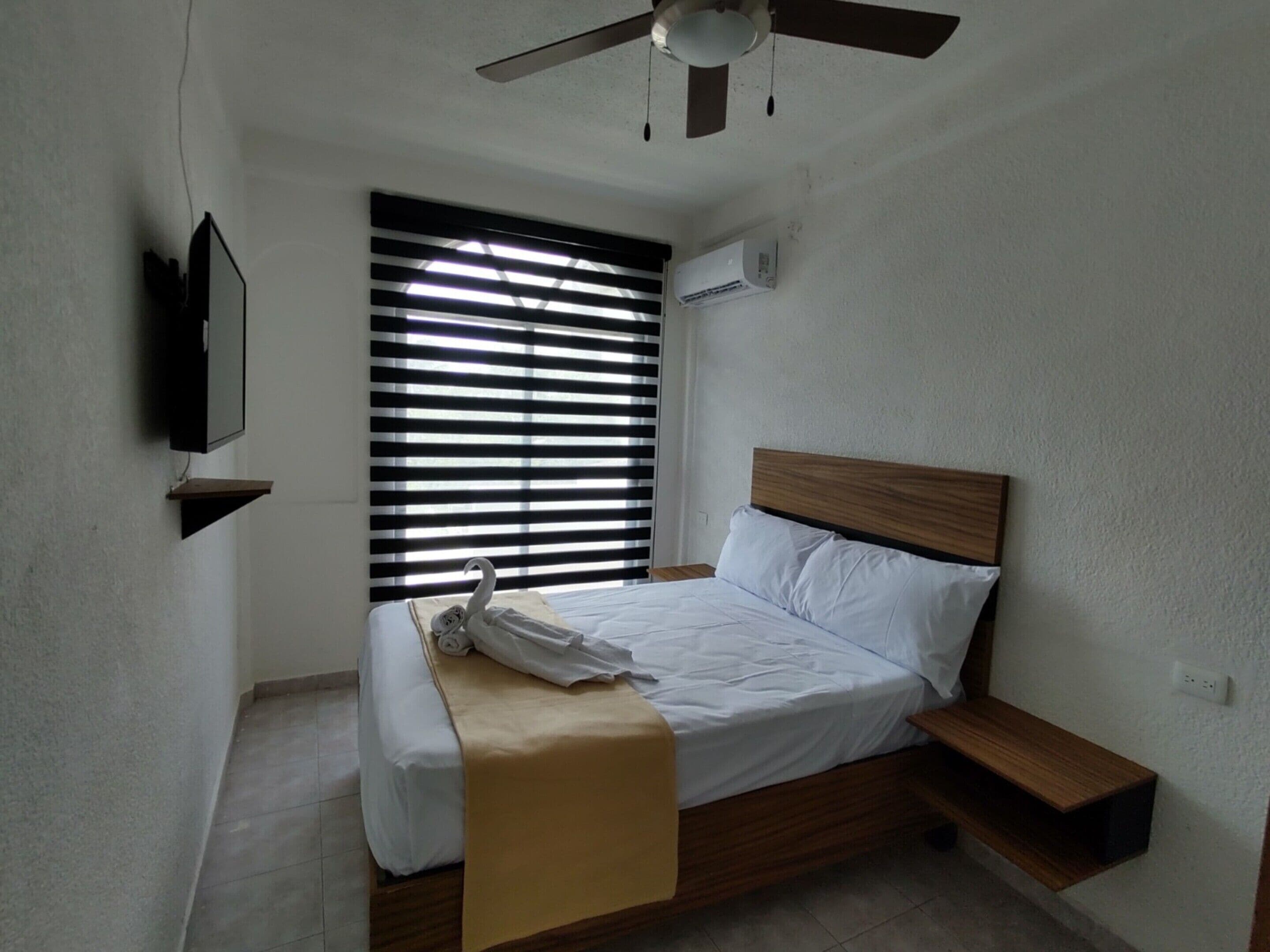 A bedroom with a bed, a ceiling fan, a window with blinds, and a mounted TV.