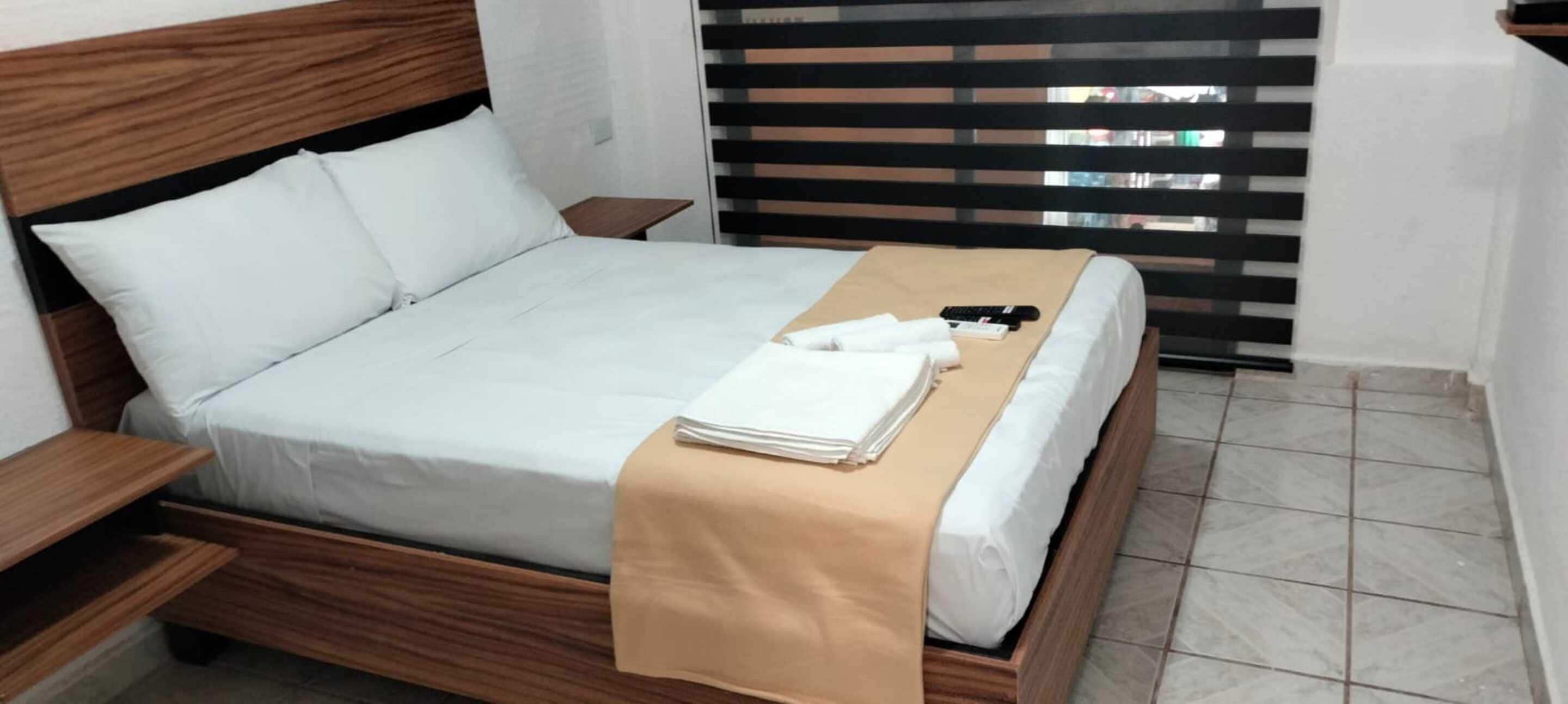 A neatly made bed with a beige blanket, white pillows, and a remote control on a wooden headboard.