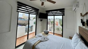 A bedroom with a bed, a ceiling fan, a TV, and a balcony with a view of buildings and a thatched roof structure. - MAYAB Playa - 5Th. Ave (Playa del Carmen)