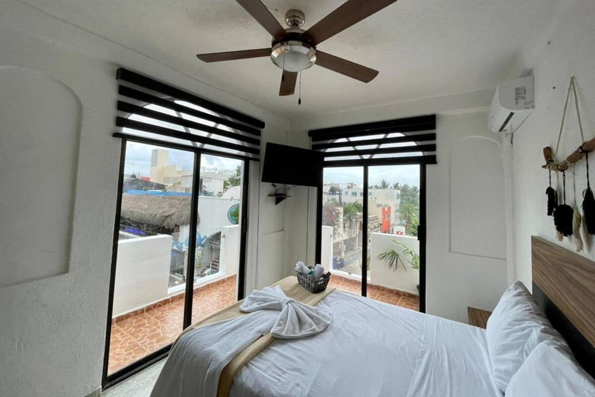A bedroom with a bed, a ceiling fan, a TV, and a balcony with a view of buildings and a thatched roof structure.