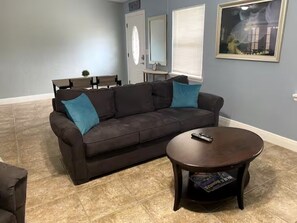 A living room with a dark brown sofa, two teal cushions, a round wooden coffee table, and a remote control on it. - Charming 3-bedroom home away from home. (Chuluota)