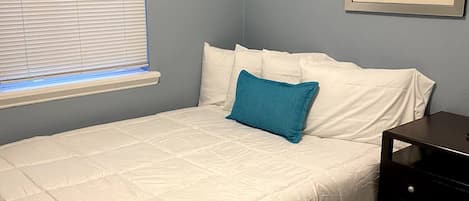 A bedroom with a bed, a window with blinds, a nightstand, and a painting on the wall.