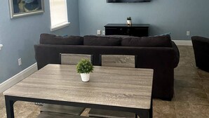 Una sala de estar con una pared azul oscuro, un televisor de pantalla plana montado sobre un mueble negro, un sofá seccional marrón oscuro, una mesa de centro de madera con una planta pequeña y una mesa de comedor con sillas.