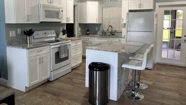 Una cocina con armarios blancos, un bote de basura de acero inoxidable, un microondas, un horno y un refrigerador.