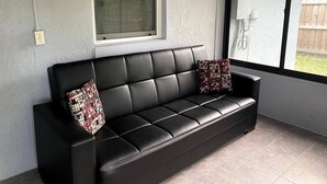 Un sofá de cuero negro con dos cojines estampados, una ventana con persianas y vista a un patio trasero con cerca de madera.