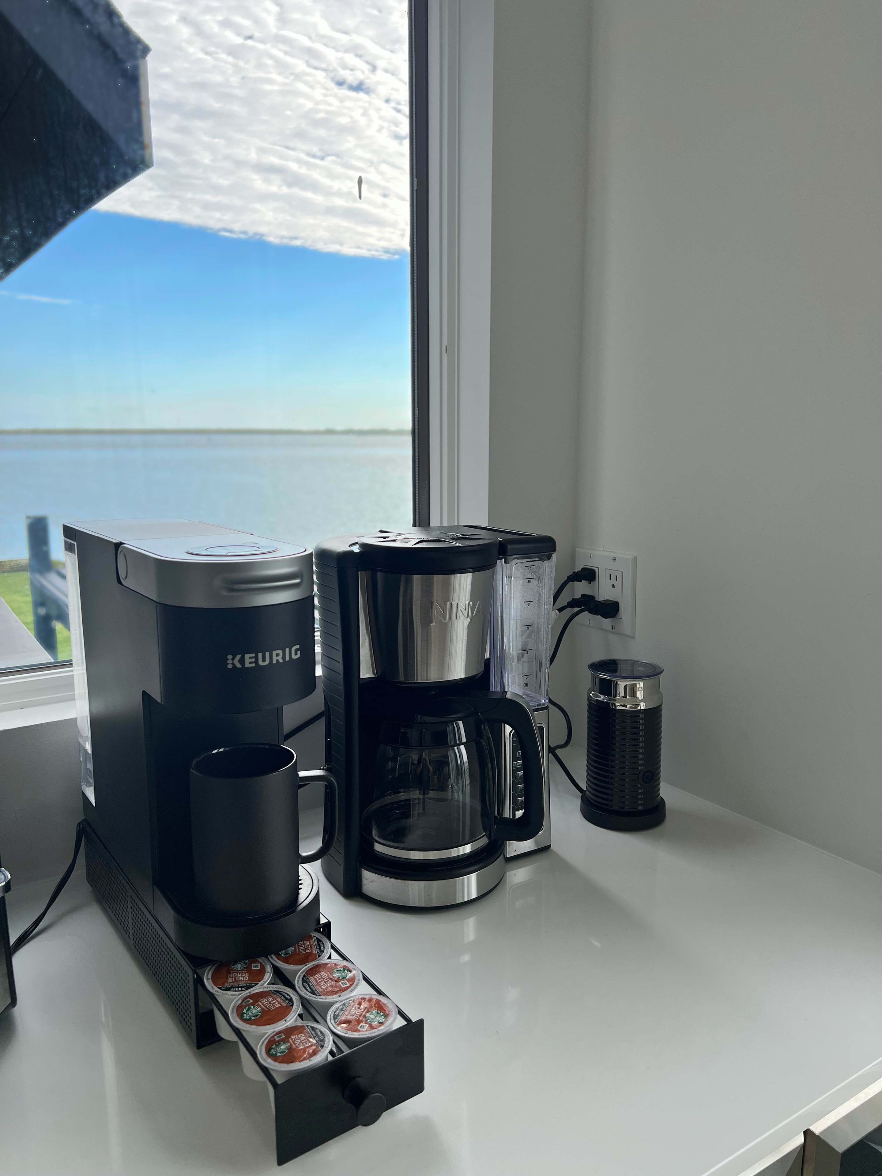 A Keurig coffee maker on a white countertop with a view of the sky and water outside.