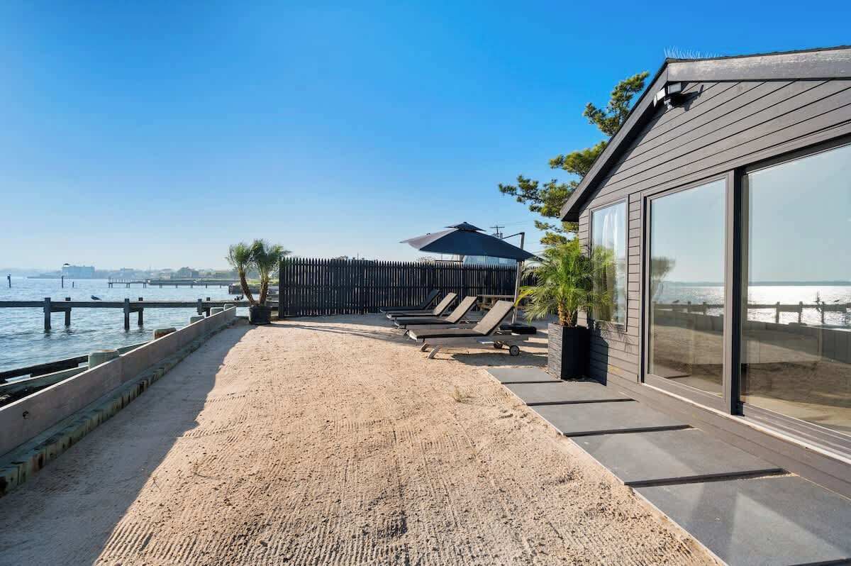 Un paseo marítimo frente a la playa con una terraza de madera, sillas de descanso y vista al agua.