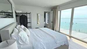 A modern bedroom with a large bed, a wardrobe, a mirror, and a view of the sea. - Black Swan Beachfront Home with Jacuzzi year round (Hampton Bays)