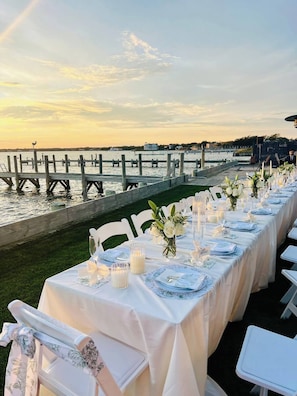 An outdoor dining area with tables set for a meal, overlooking a body of water and a dock. - Black Swan Beachfront Home with Jacuzzi year round (Hampton Bays)