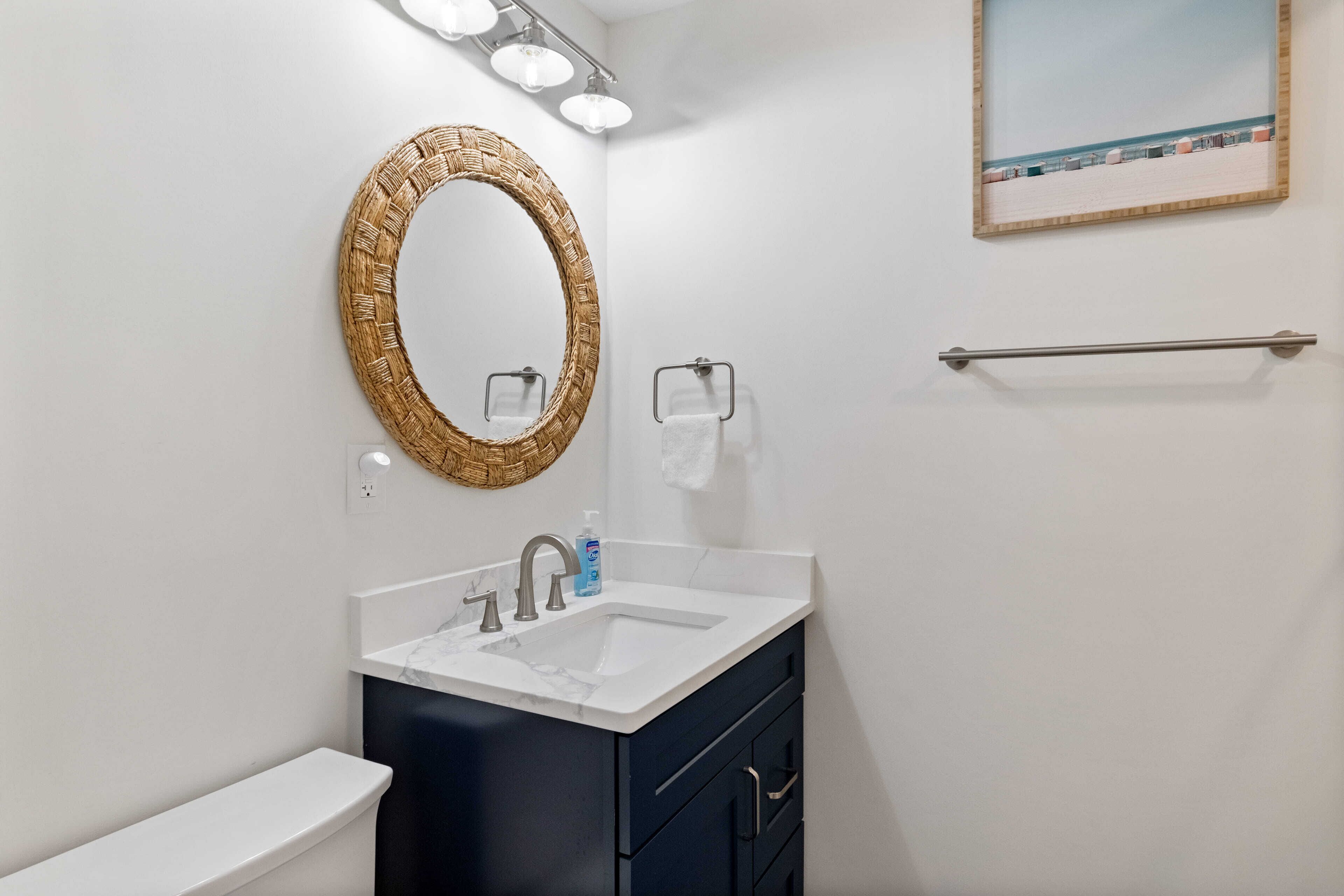 A bathroom with a white toilet, a dark blue vanity with a white sink, a woven mirror, and a towel rack.