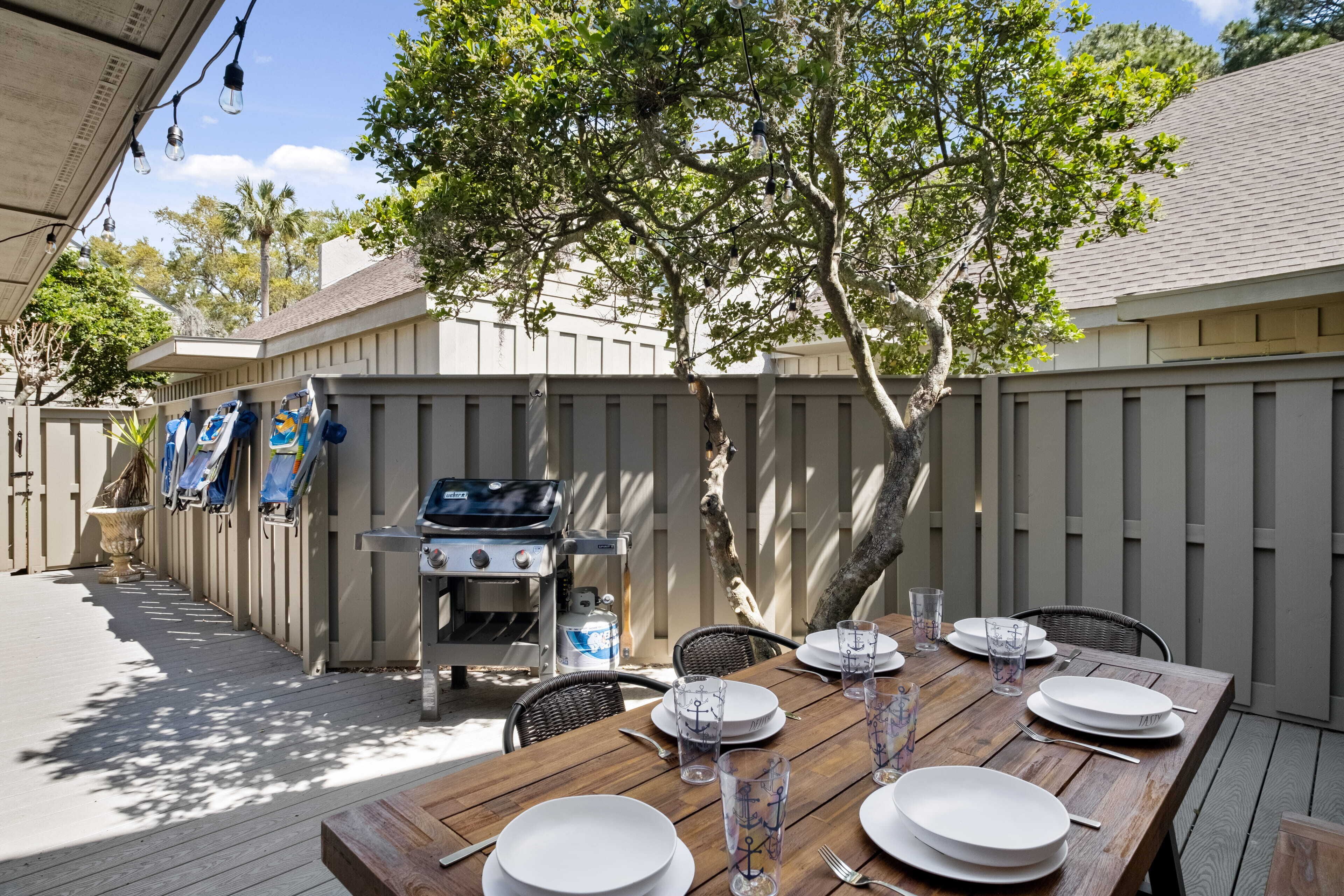 Een eethoek buiten met een houten tafel voor vier personen, omringd door een houten hek, een barbecue en een boom die schaduw geeft.