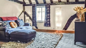A cozy attic bedroom with exposed wooden beams, a blue sofa, a matching ottoman, and a large window with curtains. - 1 bedroom accommodation in Abingdon (Abingdon)
