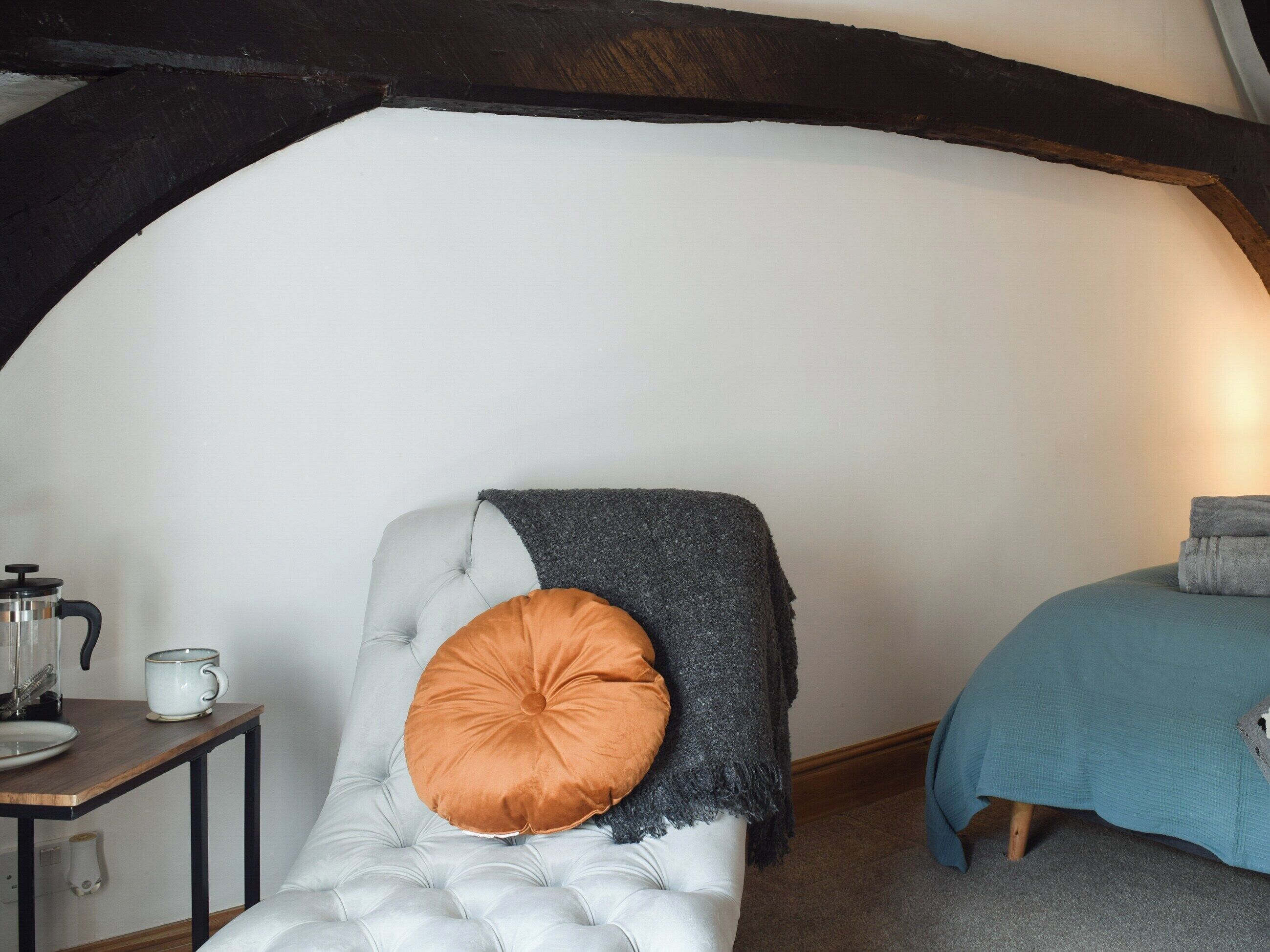 Una habitación acogedora con un sillón capitoné, una mesita con cafetera y una cama con manta azul.