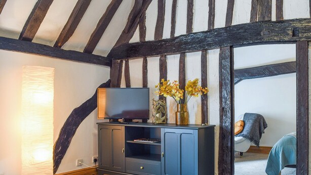 A room with exposed wooden beams, a dark blue entertainment unit, a flat-screen TV, and a vase with yellow flowers. - 1 bedroom accommodation in Abingdon (Abingdon)