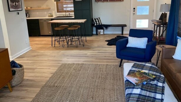 Ein modernes Wohnzimmer mit blauem Sessel, einer Couch, einem Esstisch mit vier Stühlen, einer Küchenzeile mit Fenster und einem kleinen Teppich.