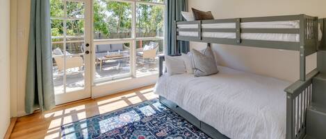 A room with a bunk bed, a large window with curtains, a patterned rug, and a view of the outdoors.