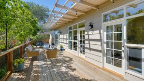 Eine Holzterrasse mit Korbmöbeln und Topfpflanzen, großen Fenstern und einer Pergola mit Lichterketten.
