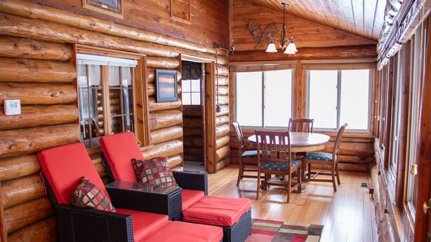 A log cabin interior with a dining area, red cushioned seating, and large windows.