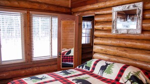 A log cabin room with a bed featuring a checkered blanket with animal prints, wooden walls, and a mirror on the wall.