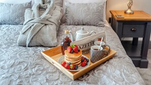Un lit bien fait, recouvert d’une couverture à motifs gris, une table de chevet avec une lampe, et un plateau avec des crêpes, du sirop et des fruits rouges.