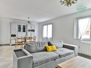 A modern living room with a grey sofa, yellow pillow, and a wooden coffee table. A dining area with a white table and chairs is visible in the background.