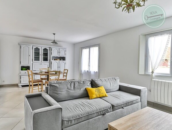 A modern living room with a grey sofa, yellow pillow, and a wooden coffee table. A dining area with a white table and chairs is visible in the background.