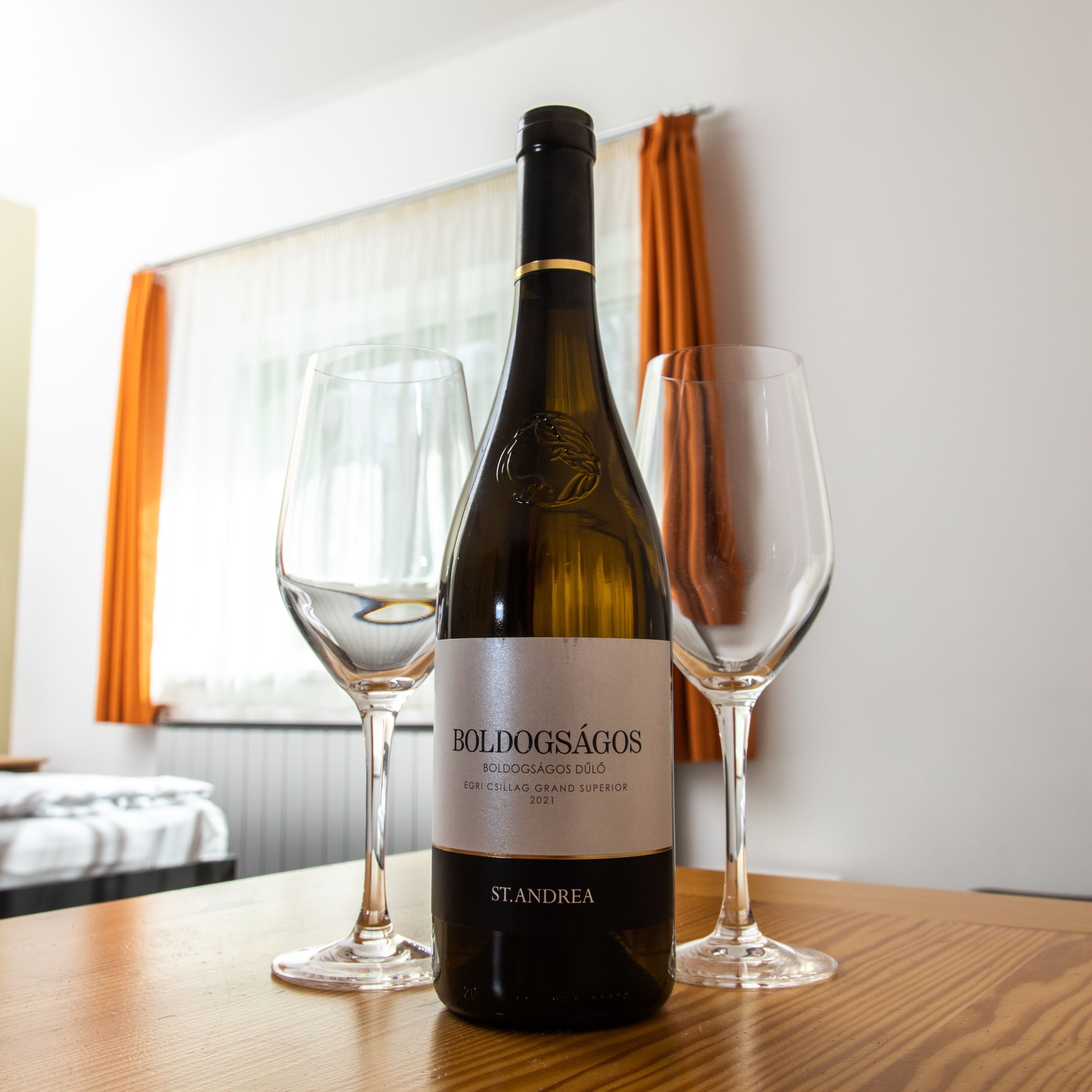 A bottle of Boldogságos wine with two empty glasses on a wooden table in a room with a window and curtains.