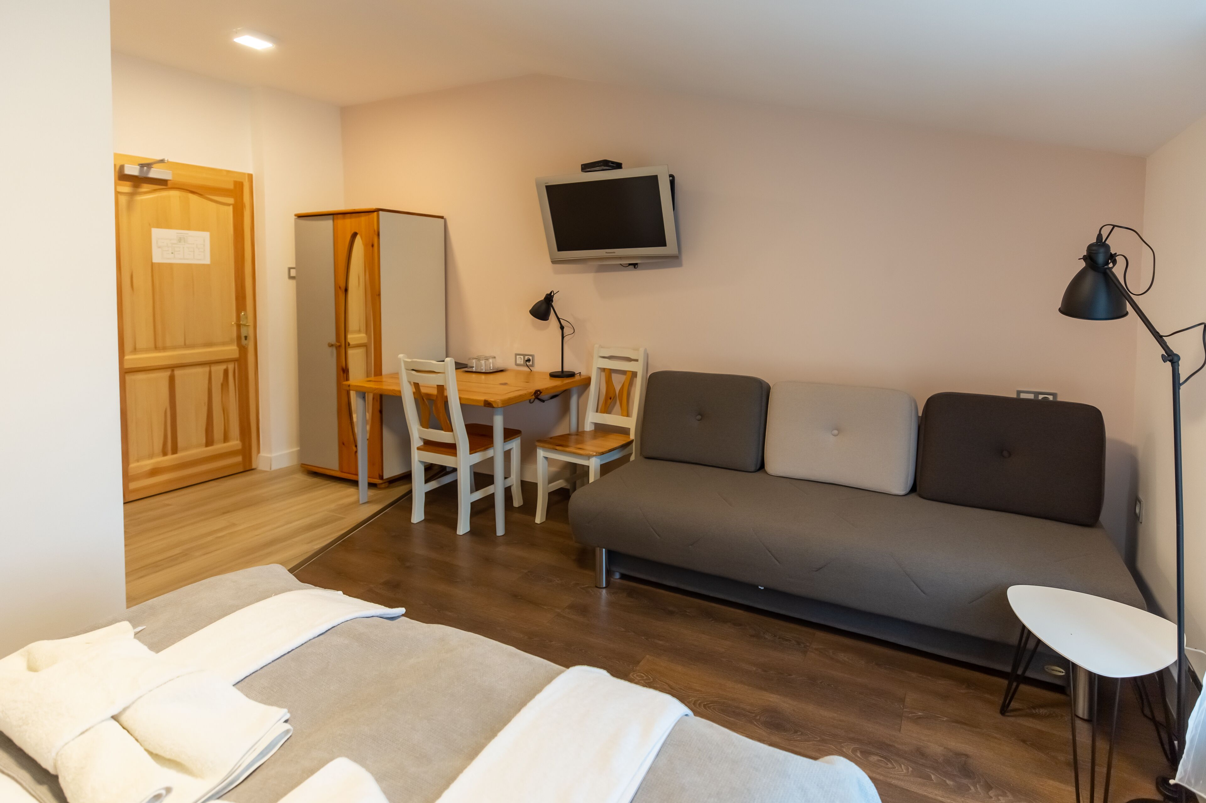 A room with a wooden door, a small table with chairs, a grey sofa, a TV mounted on the wall, and a floor lamp.