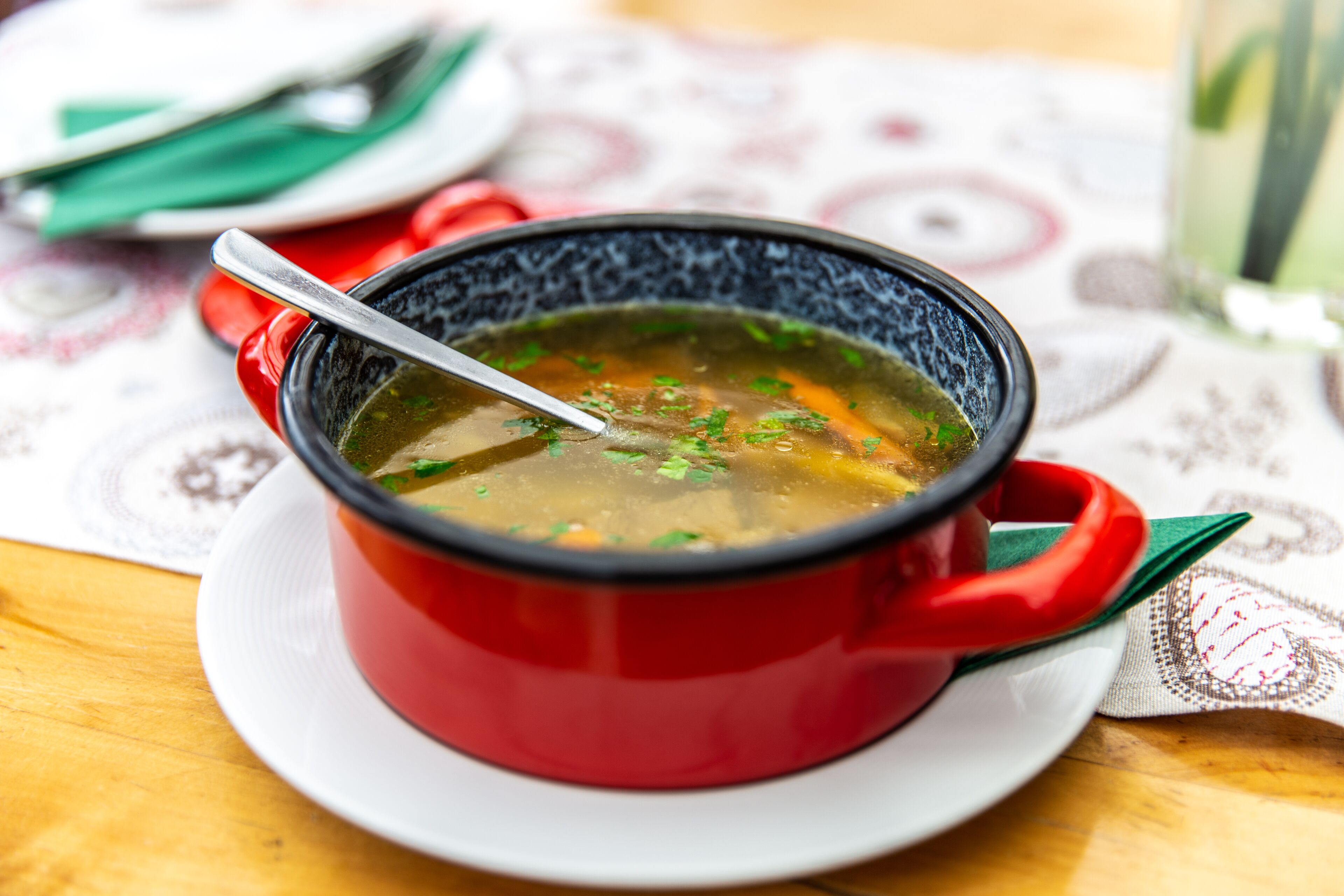 Un tazón rojo de sopa en un plato blanco, con una cuchara, sobre una mesa con mantel estampado.
