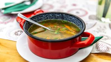 Un tazón rojo de sopa en un plato blanco, con una cuchara, sobre una mesa con mantel estampado.