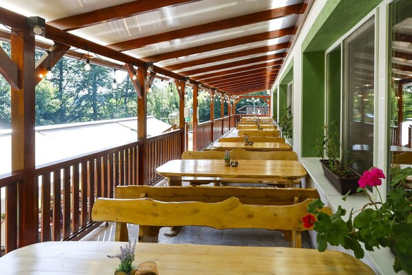 A covered outdoor seating area with wooden tables and benches, potted plants, and a view of the road. - Fehér Sas Panzió és Étterem  (Nagyvisnyó)
