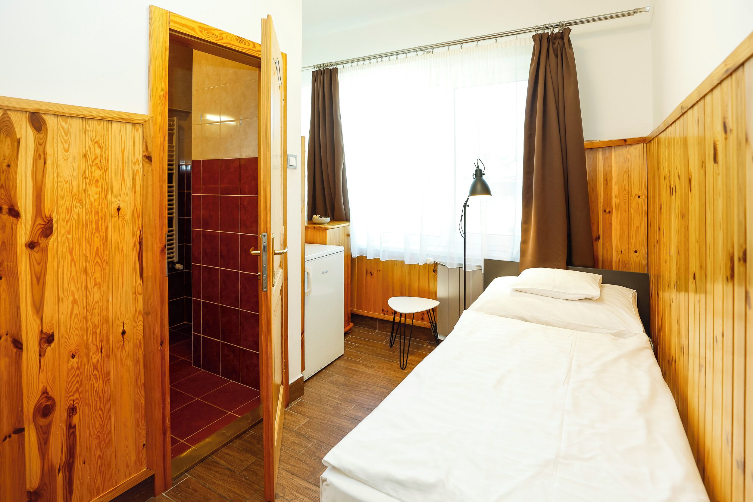 A hotel room with a wooden wardrobe, a small kitchenette, a bed with white linens, and a window with curtains.
