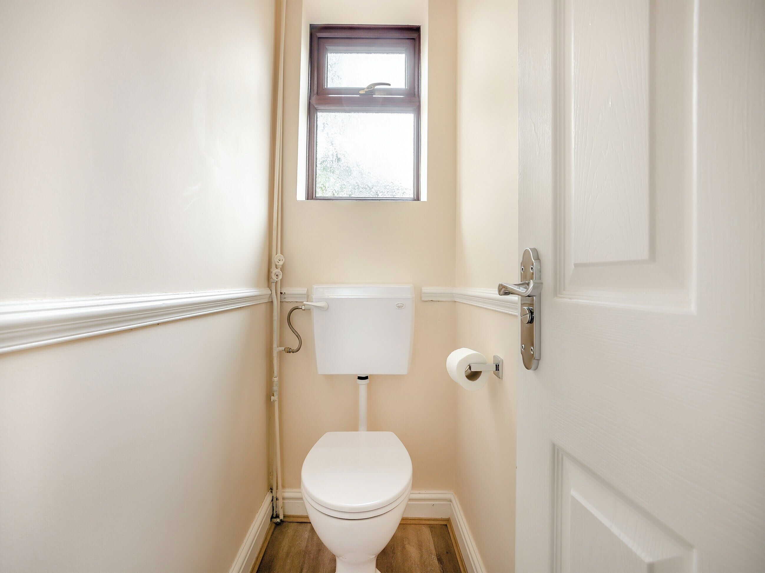 Une petite salle de bain propre, avec des toilettes blanches, une fenêtre au-dessus des toilettes et une porte dotée d’une poignée argentée.