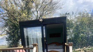 Um deck de madeira leva a uma porta de vidro que dá entrada a uma cabana com uma cama no interior.