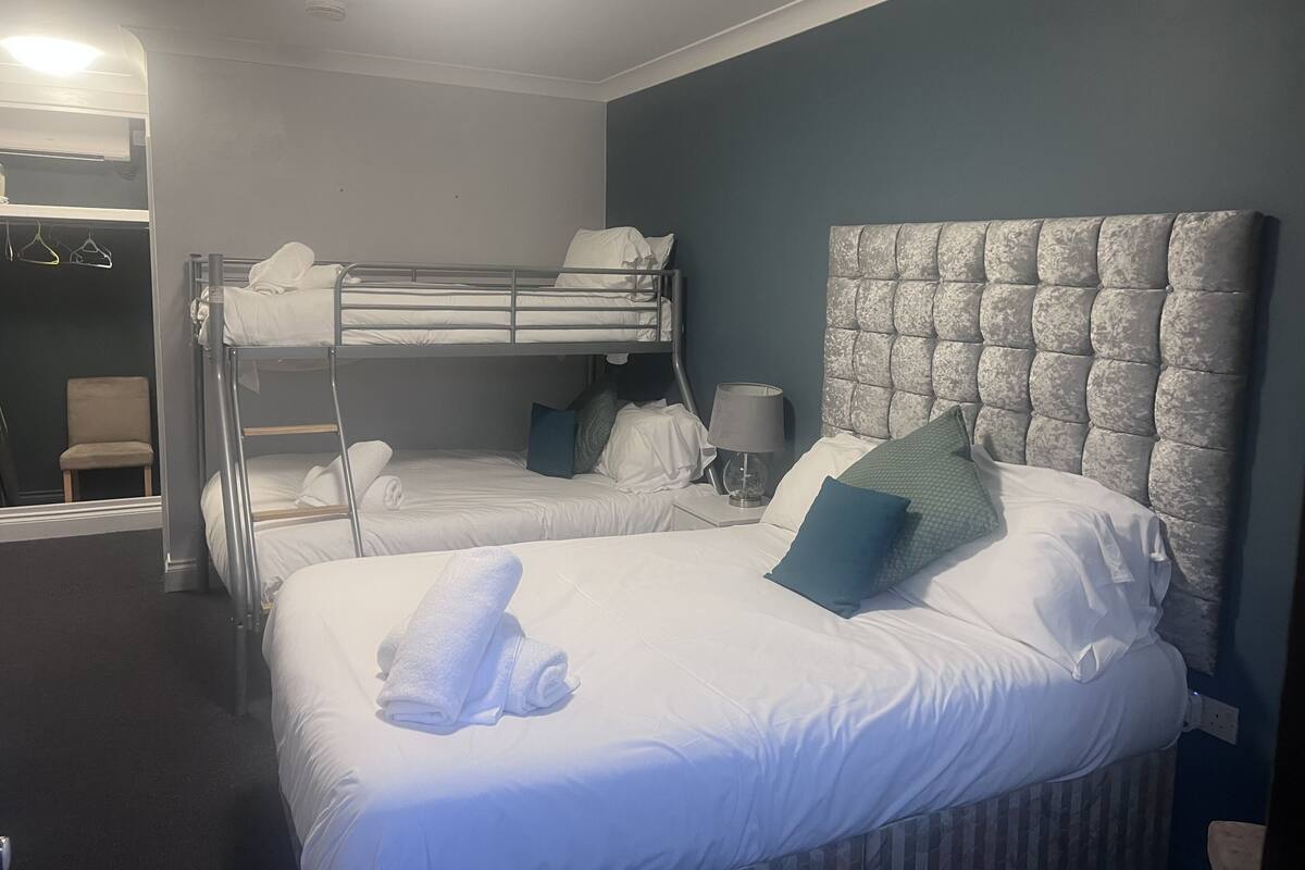 Habitació d'hotel amb un llit superposat, un llit individual, una taula de nit amb una làmpada, una cadira i un armari.