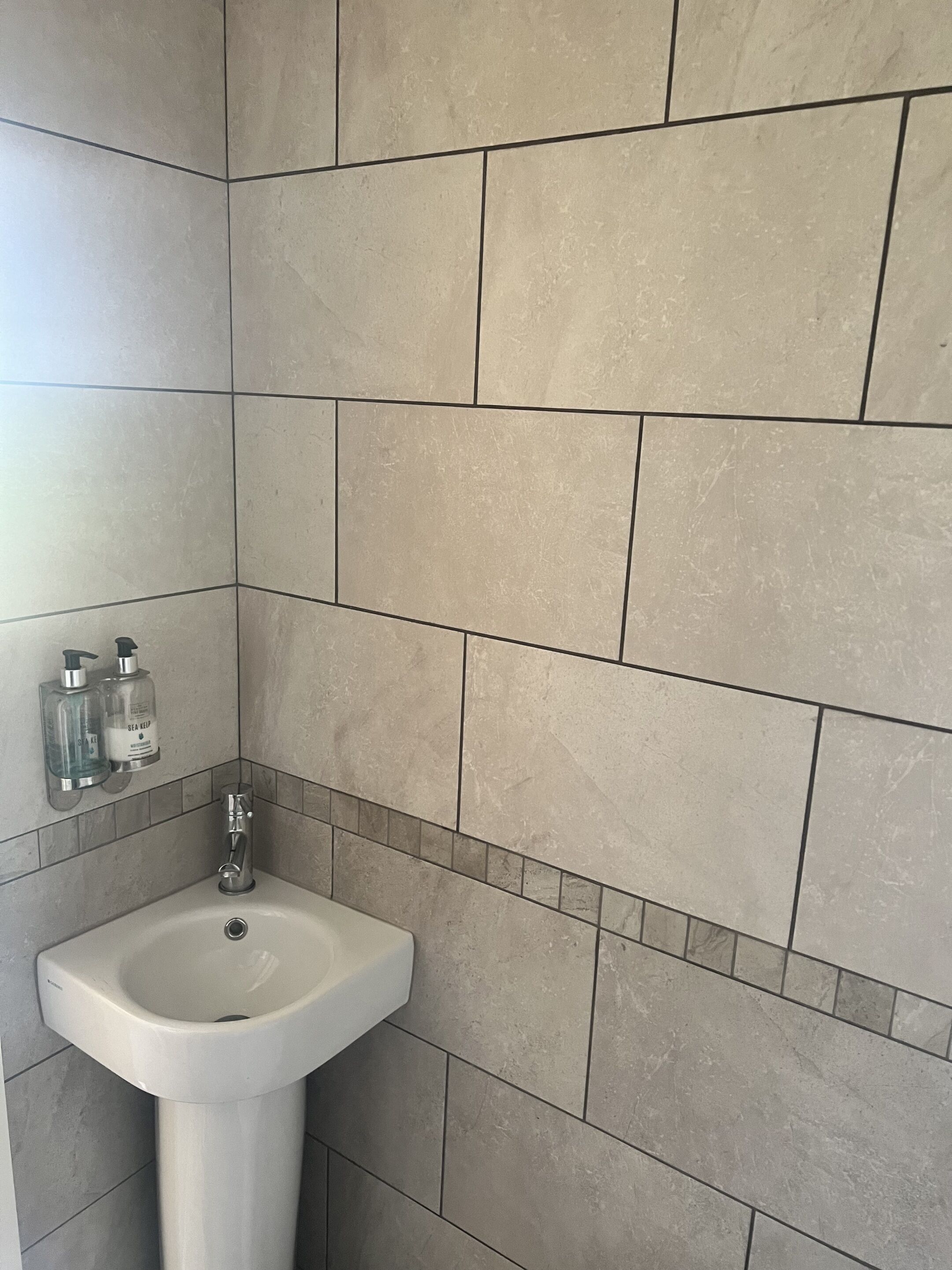 Un baño con lavamanos, dos dispensadores de jabón para manos y una zona de ducha con azulejos color beige.