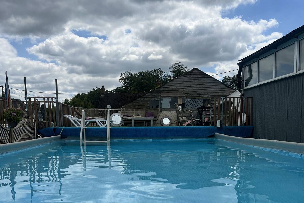 Una piscina amb paviment de rajoles, un trampolí i una zona de fusta amb cadires i una taula.