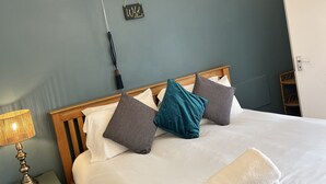 Cama bem arrumada com roupa de cama branca e cinza, um travesseiro verde-água e uma toalha branca. É visível uma cabeceira de madeira e um abajur.
